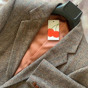 May include: A brown tweed blazer with a herringbone weave. The jacket features a peach-coloured lining and wooden buttons. A tag with two red cherries is attached to the blazer.