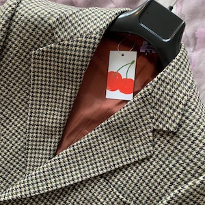 Puede incluir: Una americana con estampado de pata de gallo en marrón, negro y crema con solapa de muesca. El interior está forrado con una tela lisa de color óxido. Una etiqueta rectangular blanca con una ilustración de cerezas rojas está unida al cuello.