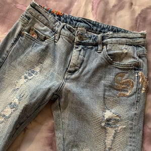 May include: Light blue denim jeans with distressed details and gold embroidery. The embroidery features a stylized letter "S" and other embellishments.