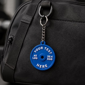 May include: A blue circular keychain shaped like a weight plate with a metal chain and ring attached to a black gym bag. The keychain features white embossed text that reads YOUR TEXT HERE, 45 LBS, and 20.4 KGS, mimicking a standard barbell plate.