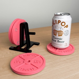May include: A set of pink circular coasters shaped like weight plates, featuring embossed text reading Standard Barbell. One coaster sits under a white aluminum can of Olipop Classic Root Beer, while two others are stored on a black stand.