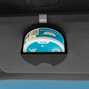 May include: A black plastic holder clipped onto a vehicle sun visor, containing a round white and teal container labeled PLUS 6 mg and 20 nicotine pouches. The holder features a semi-circular cutout at the bottom for easy access to the container.