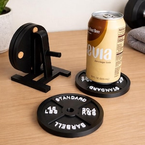 May include: Black barbell weight coasters with white lettering, one holding a can of soda. A miniature black weight machine is also present. The coasters are labeled "STANDARD BARBELL" and feature weight measurements in pounds.