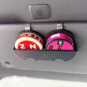 May include: A black plastic dual-slot holder clipped onto a vehicle sun visor, containing two round containers. The left container is red and white with the text ZYN 6, and the right container is bright pink and black with the text 6 MG.