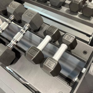 Op de afbeelding: Twee zwarte 2,27 kg dumbbells met zilveren handvatten worden getoond op een gewichtrek. De dumbbells hebben een zeshoekige vorm en het nummer "5" is aan elk uiteinde in reliëf aangebracht. Andere dumbbells zijn zichtbaar op de achtergrond.