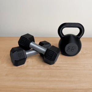 May include: A set of black dumbbells, each marked with "5", and a black kettlebell with "25" printed on it. The weights have silver handles and are arranged on a wooden surface, ideal for home workouts.