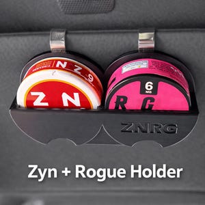 May include: A black Zyn + Rogue holder with two circular compartments, each holding a can. One can is red and white with "ZYN" and "CINNAMON" text, the other is pink with "6 MG" and "R G" text. The holder has "ZNRG" text.