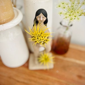 May include: A hand-painted figurine of a person with black hair and a pink face holding two yellow air plants. The figurine is standing on a wooden surface.
