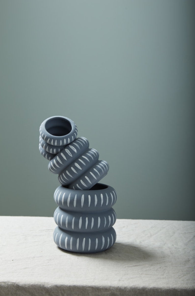 May include: A stack of four gray ceramic planters with white painted lines. The planters are stacked on top of each other and are slightly tilted to the side. The planters have a simple, modern design.