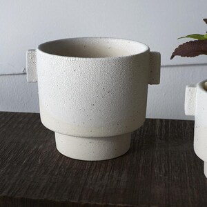 Puede incluir: Una maceta de cer&aacute;mica blanca con una superficie texturizada y agrietada. La maceta tiene forma cil&iacute;ndrica con una base escalonada y dos asas rectangulares. El color neutro y el dise&ntilde;o minimalista son adecuados para varias plantas.
