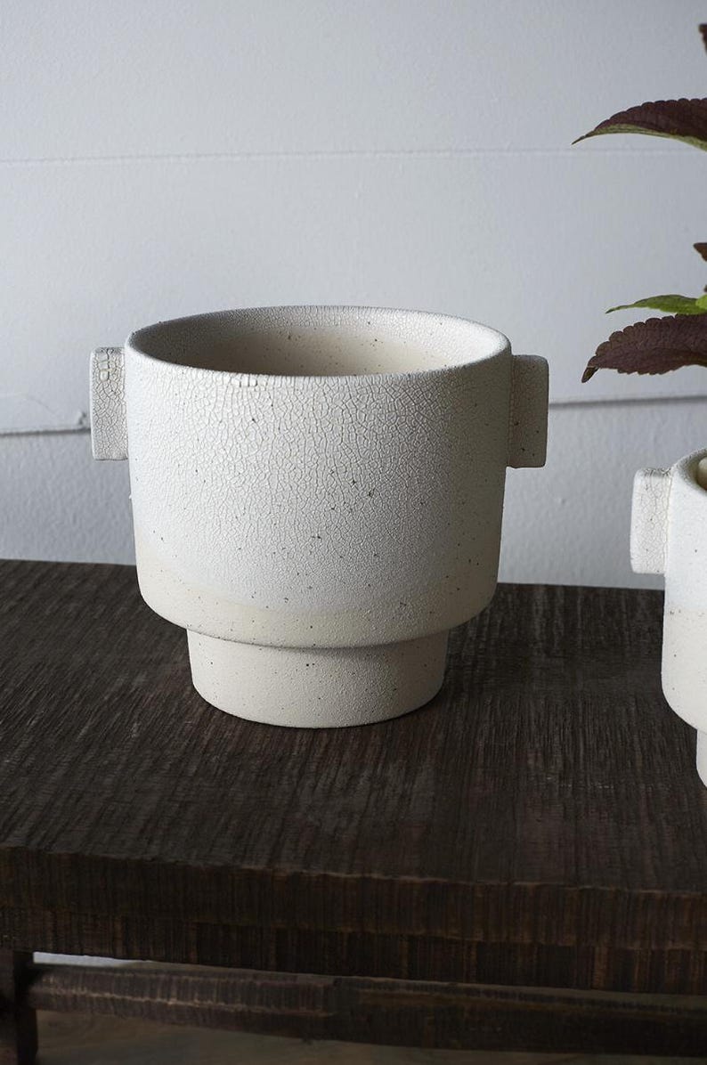Puede incluir: Una maceta de cer&aacute;mica blanca con una superficie texturizada y agrietada. La maceta tiene forma cil&iacute;ndrica con una base escalonada y dos asas rectangulares. El color neutro y el dise&ntilde;o minimalista son adecuados para varias plantas.