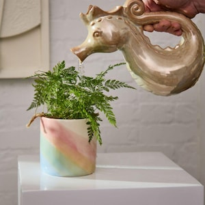 May include: A seahorse-shaped watering can pours water onto a potted fern. The ceramic watering can is tan and brown. The pot is white with pastel rainbow stripes. The plant is on a white surface.
