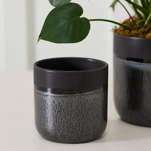 May include: Two dark gray ceramic planters. The top band is a solid dark gray, while the lower portion has a textured, speckled appearance. A green plant with large leaves is visible in the background.