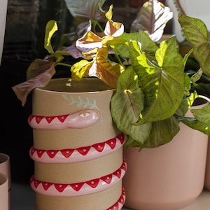 May include: A tan ceramic planter with a snake design. The snake is pink with red triangle accents. The planter is cylindrical and holds green leafy plants. Another pink planter is next to it.