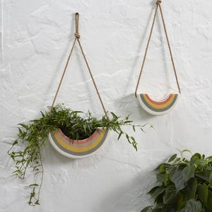 May include: Two ceramic rainbow-shaped hanging planters with rope hangers. The planters are painted with bands of color: red, orange, yellow, and gray. One planter is filled with green plants, while the other is empty.