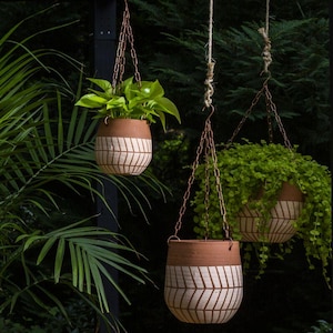 May include: Three terracotta hanging planters suspended by copper-colored metal chains and natural fiber ropes. Each pot features a smooth upper rim and a lower section decorated with a white, hand-painted chevron pattern, containing green plants.