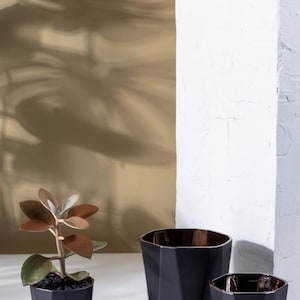 May include: Three black geometric planters with a faceted design. The smallest planter has a small succulent plant inside. The planters are on a white surface.