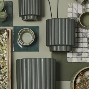 May include: A collection of various green ceramic planters with vertical ribbed detailing. The planters are in different sizes and shapes, some with plants inside. The background features decorative elements like tiles and a wire art face.