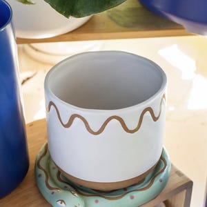 May include: White ceramic planter with a wavy gold design around the top rim. The planter sits on a light green snake-shaped saucer with pink dots. The planter is on a wooden shelf.