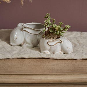 May include: Two white ceramic bunny planters with a gold trim. One planter contains a small green plant. The bunnies are lying down with their eyes closed. The planters are on a beige linen cloth.