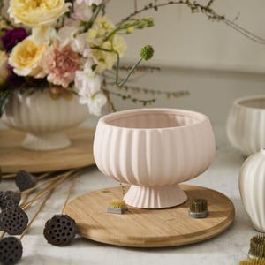 May include: A light pink ceramic vase with vertical ribbed detailing, set on a round wooden tray. The vase has a wide opening and a pedestal base. Floral arrangements and other vases are in the background. Floral arranging supplies are also visible.