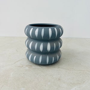 May include: A small, gray ceramic planter with white painted lines. The planter is made of three stacked rings.