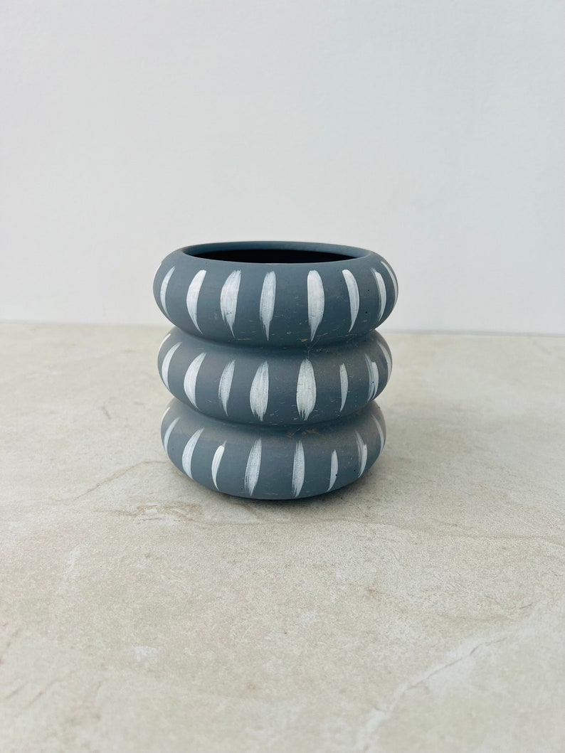 May include: A small, gray ceramic planter with white painted lines. The planter is made of three stacked rings.