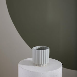 May include: A small, light gray ceramic planter with vertical ribbed detailing. The planter sits atop a white, cylindrical pedestal. The background features a muted green and off-white color scheme, creating a minimalist aesthetic.