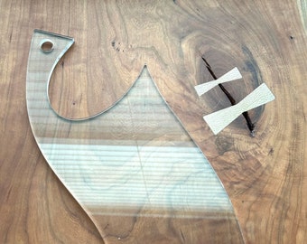 May include: A clear plastic template with a curved shape, designed for crafting or woodworking projects. The template is lying on a wooden surface.