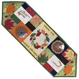 May include: A quilted Thanksgiving table runner with a green border and various square patches. The patches feature images of pumpkins, pie, leaves, and a wreath. Text reads "GIVE THANKS" and "GATHER TOGETHER".