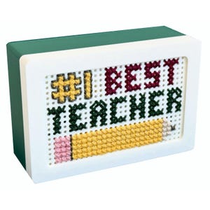 May include: A rectangular, cross-stitched plaque with the words "#1 BEST TEACHER" and a yellow pencil design. The text is in maroon, green, and yellow thread against a white background. The frame is white with a green top.