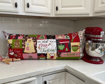 May include: A red and white Christmas themed cushion with the text "We whisk you a Merry Christmas!" and various kitchen-related images. The cushion is sitting on a white worktop next to a red stand mixer.