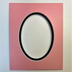 May include: A pink picture frame with a black oval mat. The mat has a white oval opening in the center.