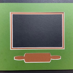 May include: A green mat with a brown and white frame around a black opening. A brown cutout of a rolling pin is below the frame.