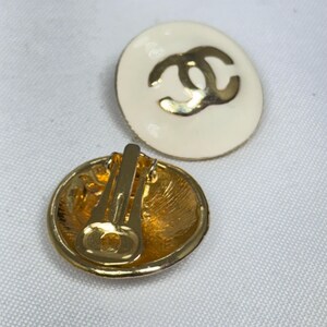 May include: Pair of clip-on earrings. Each earring is a white oval with a gold-colored double C logo. The backs of the earrings are gold-colored metal with a clip mechanism.