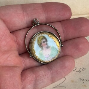 May include: A silver locket pendant with a round porcelain cameo featuring a woman in a green dress. The cameo is surrounded by a gold rim and the locket is open.