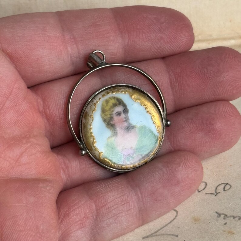 May include: A silver locket pendant with a round porcelain cameo featuring a woman in a green dress. The cameo is surrounded by a gold rim and the locket is open.