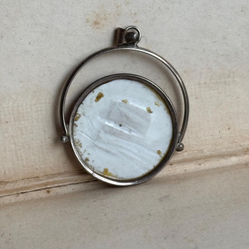 May include: A silver locket with a round, white porcelain insert. The locket has a hinged opening and a swiveling ring.
