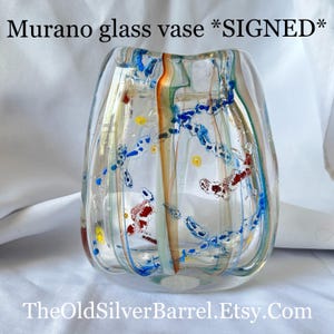 May include: Clear Murano glass vase with colorful abstract designs. The vase features blue, red, yellow, and orange accents. The text "Murano glass vase *SIGNED*" is at the top. The website address "TheOldSilverBarrel.Etsy.Com" is at the bottom.