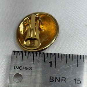 May include: Gold-toned clip-on earring with an oval shape. The earring has a textured, golden surface and a clip-on mechanism. The ruler next to the earring shows it is approximately 1 inch in diameter.