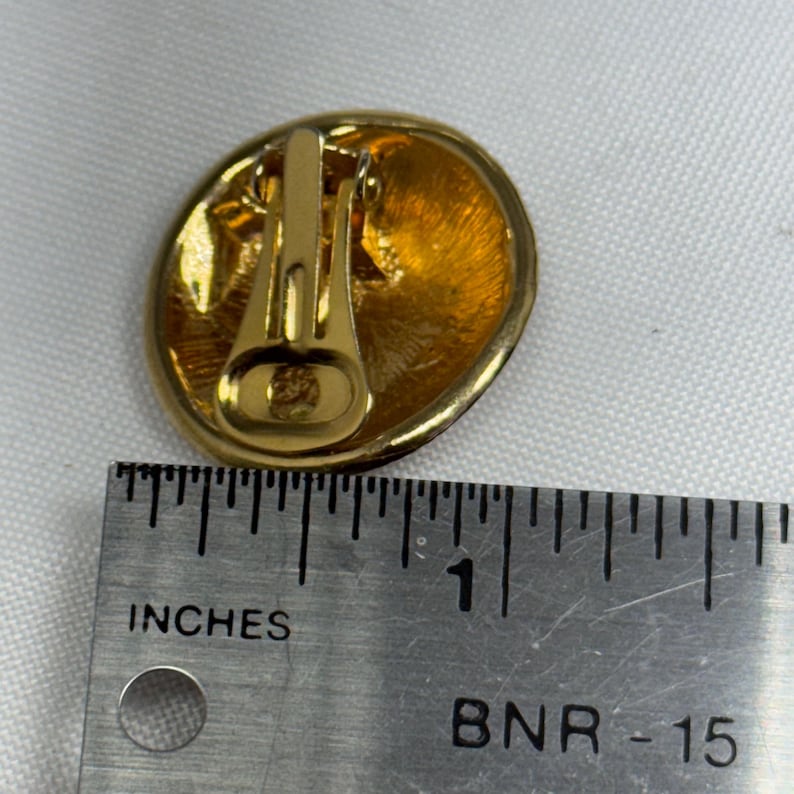 May include: Gold-toned clip-on earring with an oval shape. The earring has a textured, golden surface and a clip-on mechanism. The ruler next to the earring shows it is approximately 1 inch in diameter.