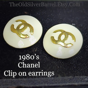 May include: Pair of vintage clip-on earrings from the 1980s. The earrings are round with a cream-colored base and a gold-colored interlocking logo. The earrings have a decorative pattern and are from Chanel.