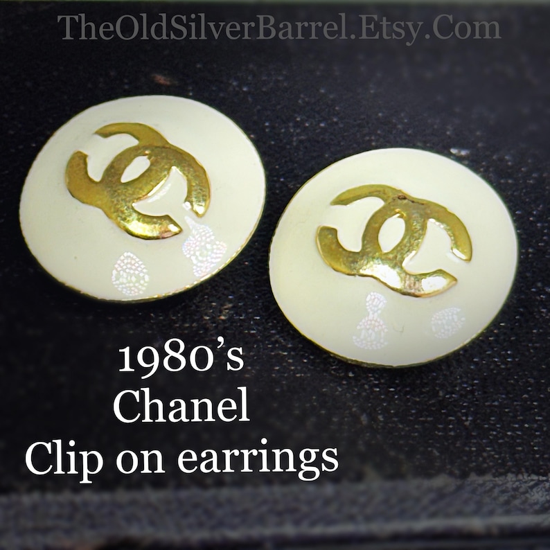 May include: Pair of vintage clip-on earrings from the 1980s. The earrings are round with a cream-colored base and a gold-colored interlocking logo. The earrings have a decorative pattern and are from Chanel.