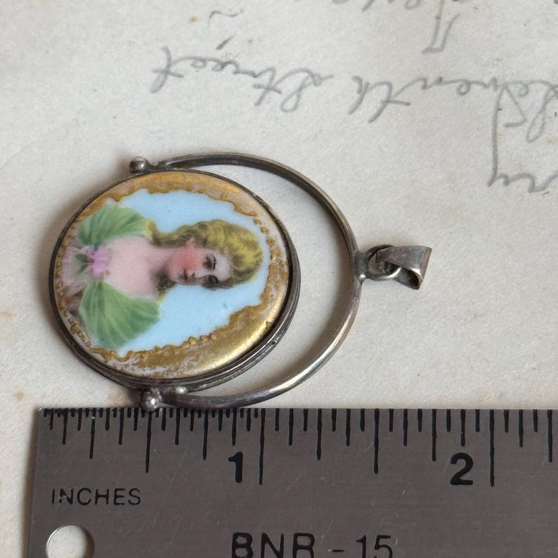 May include: A silver locket with a round porcelain cameo featuring a woman with blonde hair wearing a green dress. The locket is open and the cameo is visible. The locket is about 1 inch in diameter.