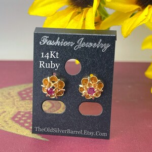 May include: A pair of 14Kt gold earrings with ruby gemstones set in a flower design. The earrings are displayed on a black card with the text "Fashion Jewelry 14Kt Ruby TheOldSilverBarrel.Etsy.Com".