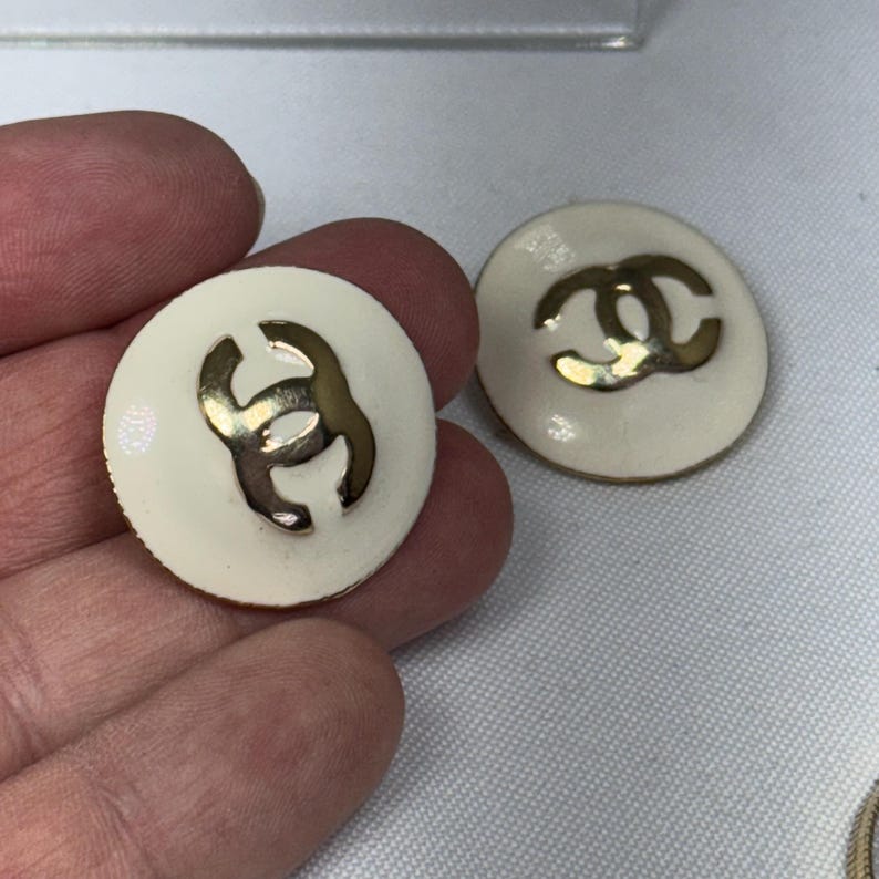 May include: Two round, off-white earrings with a gold-colored interlocking logo. The earrings have a glossy finish and a classic design. The logo is centered on each earring, creating a simple yet elegant look.
