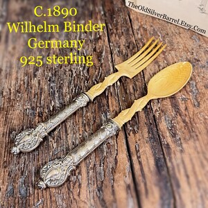 May include: Antique fork and spoon set. The set features silver handles with ornate detailing and light-colored tines and bowl. The text on the image reads "C.1890 Wilhelm Binder Germany 925 sterling". The set is displayed on a wooden surface.