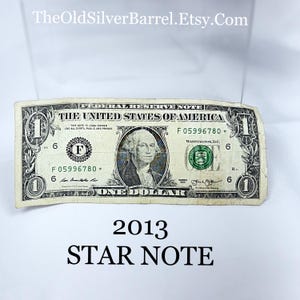 May include: A worn 2013 Star Note one-dollar bill. The bill features a portrait of George Washington and the text "The United States of America." The bill has the text "Star Note" at the bottom.