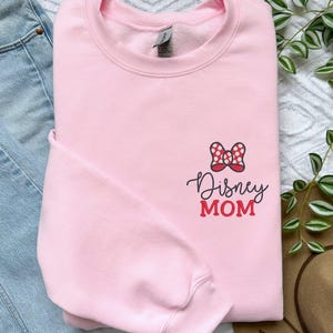 Peut inclure: Un sweat-shirt rose clair avec l'inscription "Disney MOM" en &eacute;criture noire et rouge. Un n&oelig;ud &agrave; pois rouges et blancs est plac&eacute; au-dessus du texte. Le sweat-shirt est &agrave; c&ocirc;t&eacute; d'un jean bleu et d'un chapeau marron.