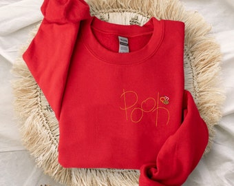 Może przedstawiać: Czerwona bluza z żółtym haftem, na którym widnieje napis "Pooh" z pszczołą obok.